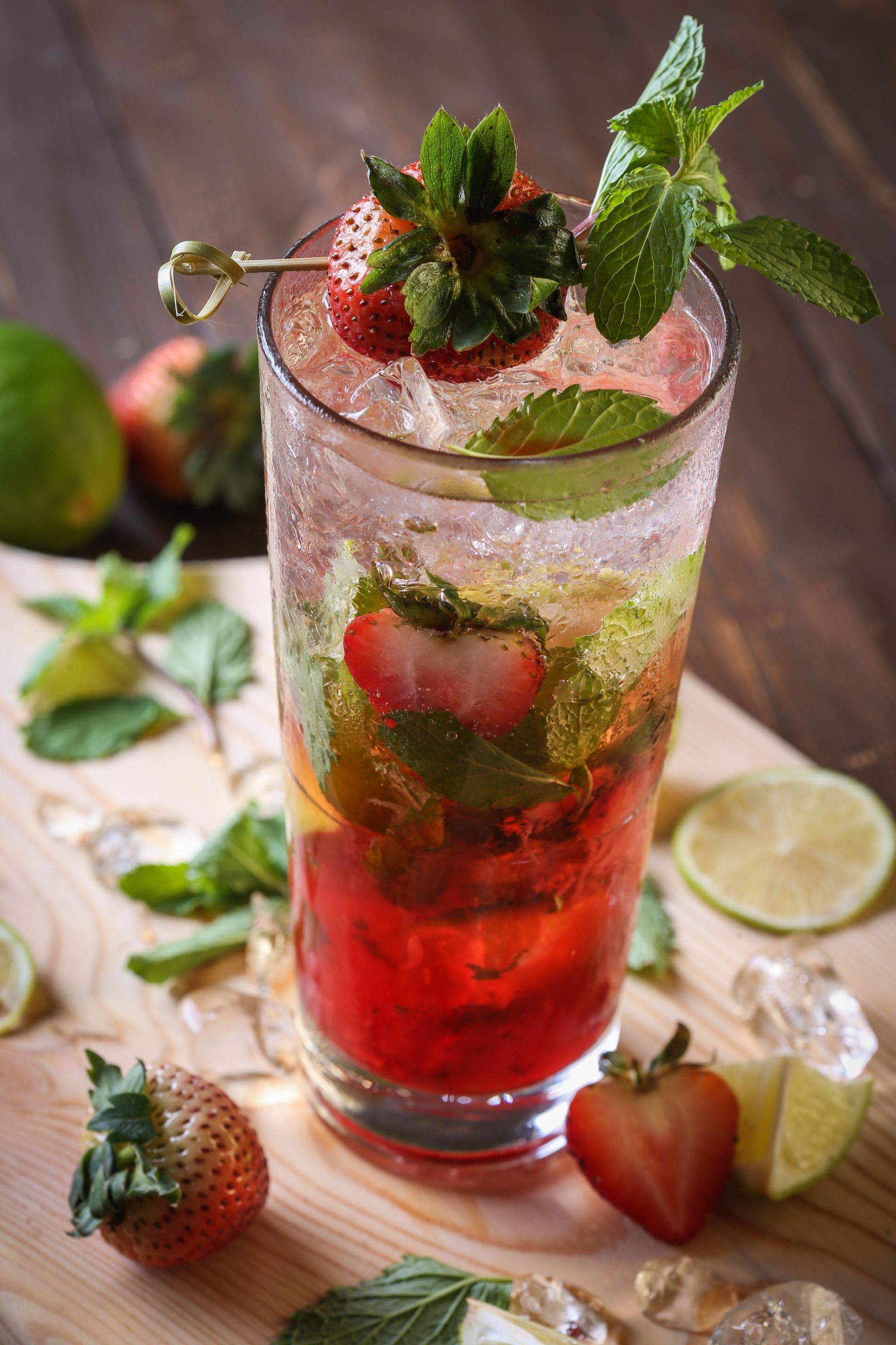 Tall glass of strawberry mint mocktail with ice and lime slices on a wooden table.